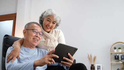 senior couple sitting on sofa using tablet while video call online with family in living room at home