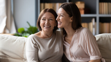 Wall Mural - Cheerful elderly mother hugs adult 30s daughter different ages and generations women laughing seated on couch at home, enjoying communication, remembering past funny stories, sharing news, feels happy