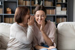 © fizkes - Middle-aged mother telling on ear to adult daughter secret, different generation women sit on sofa in living room share private hidden info laughing feels overjoyed, funny spend time together at home