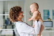 © AnnaStills - Friendly pediatrician holding baby on arms and smiling, she examining her during medical exam at hospital