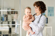© AnnaStills - Smiling doctor in white coat holding baby on arms while standing at office