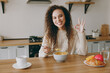 © ViDi Studio - Young woman of African American ethnicity wear casual clothes sweater eat breakfast muesli cereals with milk fruit in bowl show ok sit at table in kitchen at home alone Lifestyle cooking food concept