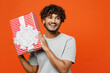 canvas print picture - Young smiling happy Indian man he wearing t-shirt casual clothes hold present box with gift ribbon bow looking aside on area isolated on orange red color background studio portrait. Lifestyle concept.