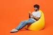 © ViDi Studio - Full body young smiling happy Indian man he wears t-shirt casual clothes sit in bag chair use mobile cell phone type message isolated on orange red color background studio portrait. Lifestyle concept.