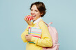 © ViDi Studio - Side view young woman student wear casual clothes yellow sweater backpack bag hold books notebooks eat apple wink blink eye isolated on plain blue background. High school university college concept