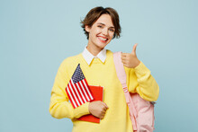 Young Smiling Happy Woman Student Wear Casual Clothes Yellow Sweater Backpack Bag Hold Notebook American Flag Show Thumb Up Isolated On Plain Blue Background. High School University College Concept.