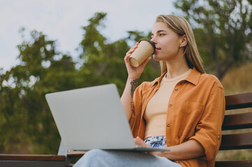 Wall Mural - Young IT woman wear orange shirt casual clothes hold use work on laptop pc computer drinking coffee sit on bench walk rest relax in spring city park outdoors on nature Urban lifestyle leisure concept