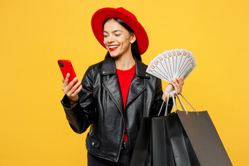 Wall Mural - Young woman wear casual clothes red hat hold shopping paper package bags fan of cash money in dollar banknotes mobile cell phone isolated on plain yellow background. Black Friday sale buy day concept.