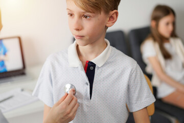 Wall Mural - Doctor and kid patient are in the clinic. Physician in white coat examining a serious young boy with a stethoscope, close up. Medicine, therapy concept