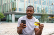 © Юлия Завалишина - African American man shopping online holding smartphone paying with gold credit card Guy on urban street in city buying on Internet enter credit card details Online shopping ecommerce delivery service