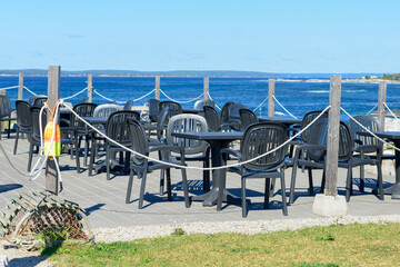 Multiple empty black plastic tables with square glass tops and black chairs at a sidewalk cafe patio of a restaurant. The outdoor restaurant has a blue and white wooden exterior with large windows.