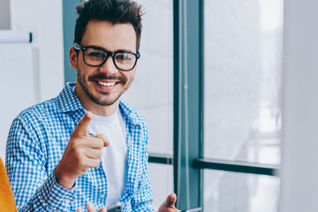 Wall Mural - portrait of cheerful millennial hipster guy in optical spectacles pointing while smiling at camera