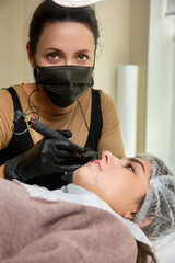 Wall Mural - A permanent makeup artist in a black protective mask on his face looks at the camera, performing lip tattooing for a young pretty woman. Make-up artist gives permanent makeup to a girl on her lips