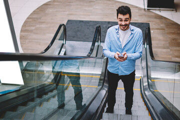 Wall Mural - portrait of cheerful hipster guy standing on escalator with smartphone gadget in hands