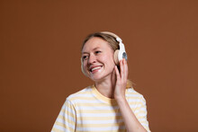 Smiling Blond Woman Listening Music Through Wireless Headphones Over Brown Background