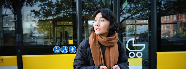 Portrait of korean girl looking for her bus on a stop, holding mobile phone, checking schedule, time table on smartphone app