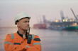 Wind Farm Offshore Maintenance Technician. Seafarer. Seaman. Navigator. A Man In A Working Overall Boiler Suit With A Radio And Safety Helmet With A Blurred Container Vessel In The Background