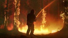 Experienced Firefighter Extinguishing A Wildland Fire Deep In The Woods. Professional In Safety Uniform And Helmet Spraying Water To Fight Large Flames Spreading Through Trees