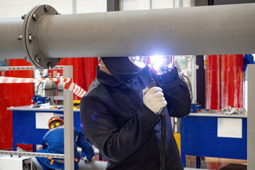 Poster - Welder repairs pipe. Man repairs metal pipeline. Guy with welding machine near pipe. Welder in production of metal pipes. Man wearing mask to protect eyes from sparks. Welder at work.