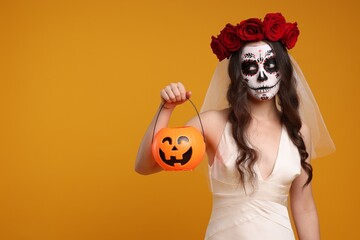 Wall Mural - Young woman in scary bride costume with sugar skull makeup, flower crown and pumpkin bucket on orange background, space for text. Halloween celebration