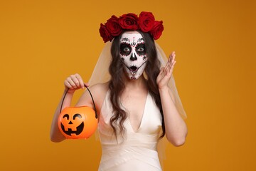 Wall Mural - Emotional young woman in scary bride costume with sugar skull makeup, flower crown and pumpkin bucket on orange background. Halloween celebration