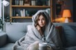 A cold woman, who has caught a cold and is suffering from the flu, is sitting on a sofa in the living room of her house. With a cup wrapped in a blanket, trying to find relief.