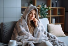 A Cold Woman, Who Has Caught A Cold And Is Suffering From The Flu, Is Sitting On A Sofa In The Living Room Of Her House. With A Cup Wrapped In A Blanket, Trying To Find Relief.