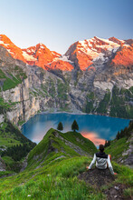 View Of A Hiker Resting In Front Of The Oeschinensee Lake Surrounded By Snowy Peak At Sunset, Oeschinensee, Kandersteg, Bern Canton, Switzerland
