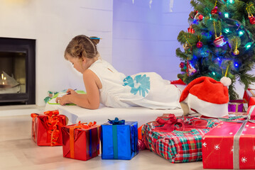 Wall Mural - Pretty little girl with the Christmas tree and gifts in front of her making a drawing for Santa