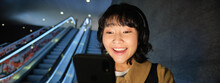 Close Up Of Happy Young Asian Girl, Looks At Her Smartphone With Surprised, Excited Face Expression, Reading Good News On Phone, Standing On Escalator In City