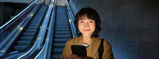 Wall Mural - Young Japanese girl standing with smartphone, going down escalator with mobile phone, commuting in city, using public transport