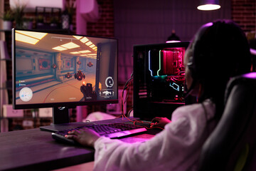 Woman in cozy apartment playing captivating video game on gaming PC at computer desk late at night. Sleepy gamer girl battling villains in multiplayer science fiction shooter