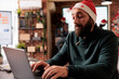 © DC Studio - Startup company worker typing message on laptop while working in office at christmas eve. Corporate employee wearing santa claus hat using computer and sitting at workplace desk