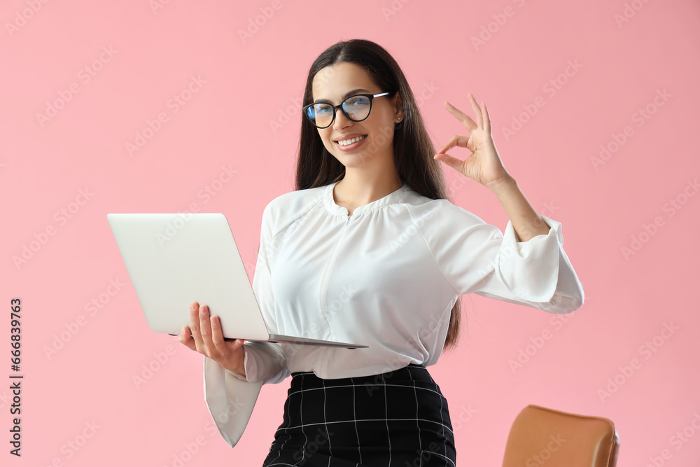 Pretty young female programmer with laptop showing OK gesture on pink background