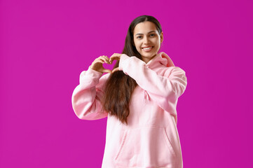 Wall Mural - Young woman in pink hoodie making heart gesture on purple background