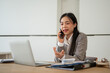 © bongkarn - A professional Asian businesswoman is talking on the phone with her client at her office desk.
