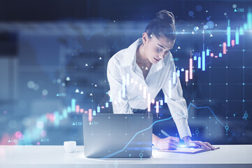 Poster - Attractive young european businesswoman writing in notepad while leaning on desk with laptop and glowing forex chart on blurry office interior background. Trade and market concept. Double exposure.