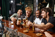group of four happy multiethnic male friends toasting with glasses of beer at bar counter