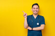 © Bangun Stock Photo - Smiling professional young Asian male doctor or nurse wearing a blue uniform standing confident while pointing a finger at copy space isolated on yellow background. Healthcare medicine concept