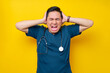 Stressed professional young Asian male doctor or nurse wearing a blue uniform holding a head and screaming isolated on yellow background. Healthcare medicine concept