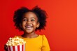 excited movie, portrait smiling African American child girl eating popcorn from big cinema red striped box isolated over red background.