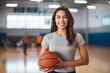Portrait of Young beautiful female basketball coach standing in school gym , holding basketball ball , looking at camera and smiling