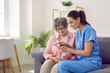 © Studio Romantic - Young pretty nurse at home shows an elderly woman a photo on a mobile phone and tells positive happy moments of life, conveying a good mood and a positive attitude. Concept of psychological health.