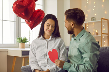 Wall Mural - Young couple in love celebrating St Valentine's Day and having a romantic date at home. Happy man and woman with a red heart-shaped Valentine card sitting on the sofa at home. Love and romance concept