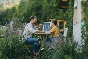 Father with daughter sitting outdoors in garden, with model of solar panel. Solar energy and sustainable lifestyle of young family. Concept of green energy and sustainable future for next generations.