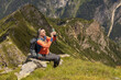 © Guzel - Gray-haired man with backpack sitting on rocky ledge and taking pictures of summer mountain landscape, Austria