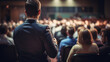 © B & G Media - A speaker giving a lecture to an audience in an auditorium, seen from behind, emphasizing the seminar's engaging atmosphere.
