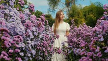 Caucasian Woman With Long Hair In White Dress Walks Among Vibrant Purple Wildflowers In Spring Field. Beautiful Nature, Blooming Flowers, Fresh Air, Beauty Concept