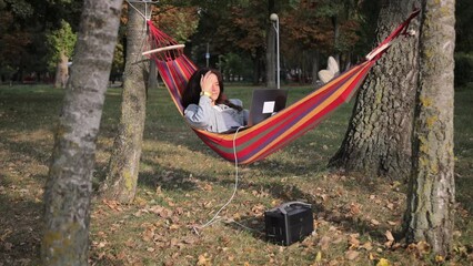 A young female programmer is working on a laptop under a tree and charging it from a portable charging station