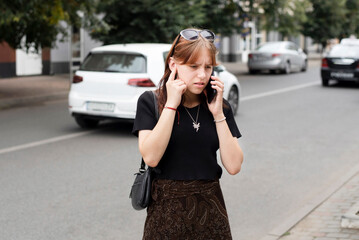 A modern teenage girl is talking on phone in the city and has to cover her ears because she can't hear her friend on the phone because of the busy noise of cars in the city